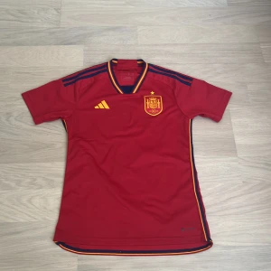 Spansk landslagströja från Adidas - Röd fotbollströja från Adidas med Spaniens landslagsmärke på bröstet. Tröjan har gula och blå detaljer, korta ärmar och rund hals med randigt mönster. Tillverkad i lätt och ventilerande material, perfekt för match eller träning. Från säsong 22/23. 