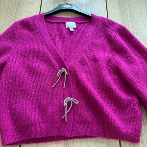 Ceriserosa stickad kofta från H&M - Säljer en ceriserosa stickad kofta från H&M med croppad passform. Koftan har långa ärmar, v-ringning och knytdetaljer framtill. Perfekt för att addera färg till din outfit!