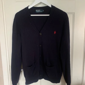 Mörkblå kofta från Polo Ralph Lauren - Klassisk mörkblå kofta från Polo Ralph Lauren med v-ringning och knäppning framtill. Tröjan har två fickor och en röd broderad logga på bröstet. Perfekt för lager-på-lager och stilrena outfits.