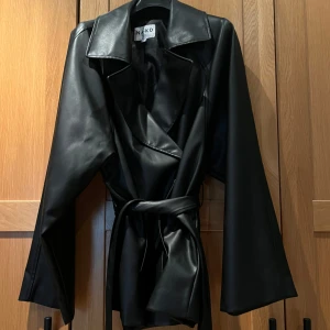 Svart skinnjacka med bälte från NA-KD - Säljer en svart skinnjacka från NA-KD med bälte i midjan. Jackan har klassisk krage och långa ärmar, perfekt för en cool och stilren look. Materialet är syntetiskt skinn och modellen är rak med markerad midja.