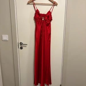 Röd långklänning med volang från Zara - Säljer en röd långklänning från Zara med smala axelband och volangdetaljer upptill. Klänningen har en liten öppning framtill och är tillverkad i ett glansigt material som ger en lyxig känsla.