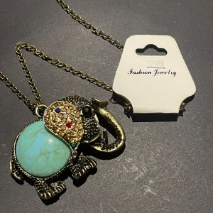 Halsband med elefantberlock och turkos sten - Hej jag säljer en elefantberlock i färg av antikguld. Elefanten har en stor turkos sten som kropp och huvudet är dekorerat med små färgglada stenar i blått, rött och grönt. Kedjan är i guldfärg och berlocken har fina detaljer. Märket är fashion jewelry som det står på etiketten