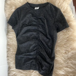 Glittrig svart topp från Kappahl - Andvänt två gånger. Säljer en glittrig svart topp från Kappahl med korta ärmar och snygg rynkdetalj framtill. Perfekt för dig som vill sticka ut lite extra med en enkel men cool look. Toppen har rund halsringning och är i stretchigt material.