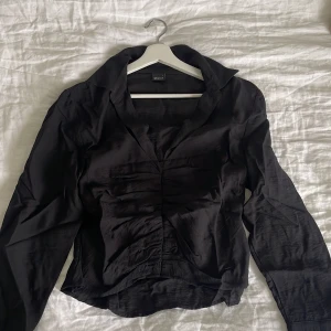 Svart långärmad skjorta från Gina Tricot - Säljer en svart långärmad skjorta från Gina Tricot med v-ringad krage och lätt rynkade detaljer framtill. Skjortan har en figurnära passform och är tillverkad i ett tunt, mjukt material som känns skönt mot huden.