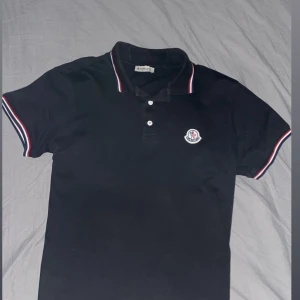 Svart piké t-shirt från Moncler - Stilren klassisk svart piké ifrån Moncler som bara används ett fåtal gånger. Med klasiska vit-röd-blå ränderna runt kragen och ärmarna, perfekt nu inför våren. Storlek S men passar M utan problem. Ps pris kan diskuteras vid snabb transaktion
