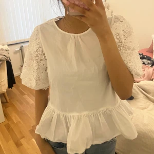 Vit Zara blus med spetsärmar och volang - Säljer en söt vit blus med korta spetsärmar och volang nertill. Ärmarna har ett gulligt blommigt spetsmönster. Blusen har rund halsringning från Zara och är helt oanvänd.
