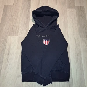 Mörkblå hoodie från GANT - Säljer en mörkblå hoodie från GANT med broderad logga och amerikanskt sköldmärke på bröstet. Tröjan har huva och magficka, samt ribbade muddar vid ärmslut och nederkant. Klassisk och stilren design.