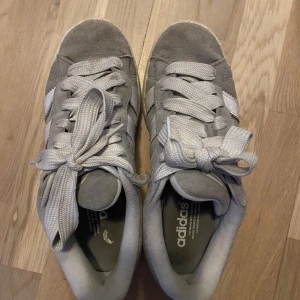 Grå Adidas Campus sneakers i mocka - Säljer ett par grå Adidas Campus sneakers i mocka med vita ränder och vit sula. Skorna har klassisk snörning och gummisula i brunt. Perfekta för dig som gillar stilrena och tidlösa sneakers. Man kan hämta upp dem fraktar inte❤️❤️