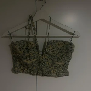Grön croppad linnetopp med mönster från Zara - Säljer en grön croppad linnetopp från Zara med smala axelband och ett detaljerat paisleymönster i gult och svart. Toppen har rynkade detaljer och knytning vid axlarna. Perfekt för varma dagar!