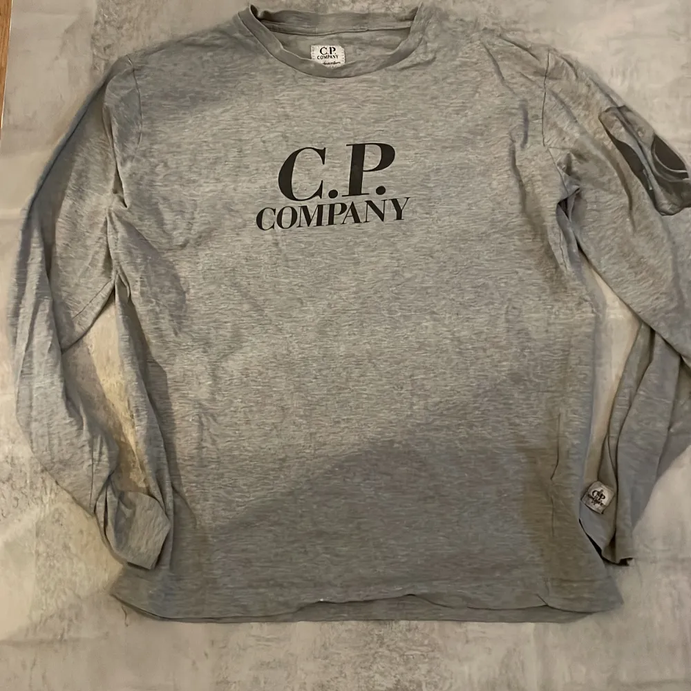 Säljer en grå långärmad tröja från C.P. Company med tryckt logga på bröstet och en unik detalj på ena ärmen. . Neuletakit & Villapaidat.