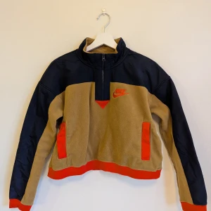 Nike - Cropped fleecetröja med magficka S - Tröja storlek S i beige fleece med marinblå ärmar och axlar samt röda detaljer. Ärmarna är i ett vindjacksliknande material på utsidan (fleece på insidan). Halvlång dragkedja framtill, röd Nike-logga på bröstet och röda muddar. En stor ficka på magen med två öppningar. Från rökfritt och djurfritt hem.