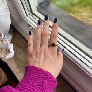 Guldfärgad ring med svart sten - Säljer en snygg guldfärgad ring med en oval svart sten i mitten. Ringen har fina detaljer på sidorna och passar perfekt som statement-smycke. Enkel men elegant design som drar blickarna till sig.