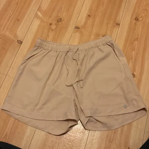 Beige shorts från Zink med dragsko - Snygga beige shorts från Zink med elastisk midja och dragsko. De har en enkel design med sidofickor och en liten metallogo framtill. Perfekta för varma dagar och en avslappnad stil. Skirt tyg. 