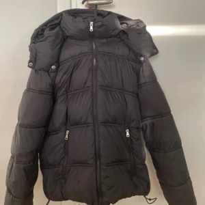 Svart pufferjacka med avtagbar huva - Säljer en svart pufferjacka med dragkedja framtill och två fickor med dragkedja. Jackan har en avtagbar huva och justerbar nederkant med snörning. Perfekt för kyligare dagar och enkel att matcha med olika outfits.