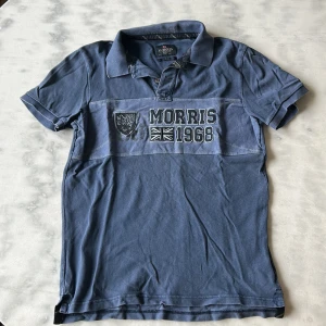 Blå pikétröja från Morris - Säljer en blå pikétröja från Morris med broderad logga, texten 'Morris 1968' och brittisk flagga på bröstet. Tröjan har klassisk krage och korta ärmar. Perfekt för en stilren och avslappnad look. Fint använt skick. Lite stonewash känsla över den. 
