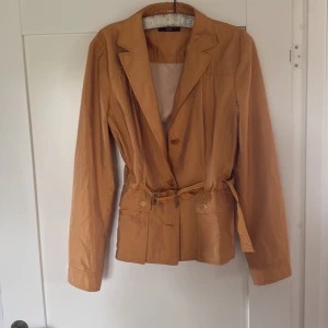 BOSS. Beige kavaj/ jacka, med bälte från BOSS - Säljer en snygg beige kavaj/ jacka från BOSS med bälte i midjan. Jackan har klassisk krage, långa ärmar och knappar framtill. Perfekt för dig som vill ha en stilren och tidlös look. Fint beige guld tyg. 