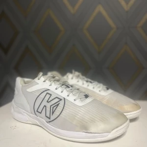 Vita sneakers från Kempa - Säljer ett par vita sneakers från Kempa med stora svarta loggor på sidorna. Skorna har snörning och en sportig design med rund tå och platt sula. Perfekta för dig som gillar stilrena och sportiga skor.
