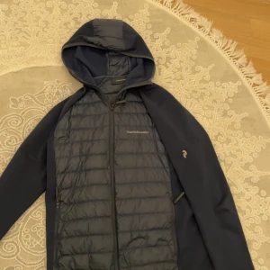 Blå hybridjacka med huva från Peak Performance - Snygg blå hybridjacka från Peak Performance med vadderad framsida och släta ärmar. Jackan har huva, dragkedja och diskret logga på bröstet. Perfekt för lager-på-lager och passar till många tillfällen. Perfekt för Våren som är här samt höst.