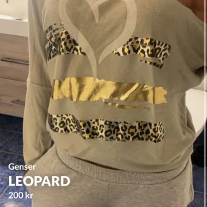 Beige tröja med leopard- och gulddetaljer - Säljer en beige långärmad tröja med leopardmönster och guldiga ränder framtill. Tröjan har en stor hjärtformad detalj på bröstet och är lite lös i passformen. Perfekt för dig som gillar djurmönster och metallic!