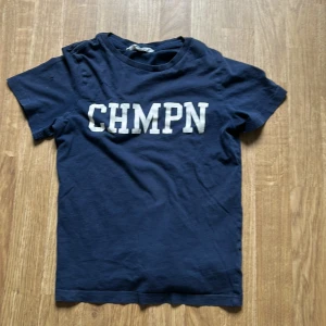 Mörkblå t-shirt med CHMPN-tryck - Mörkblå t-shirt med vit text 'CHMPN' på bröstet. Klassisk rund hals och korta ärmar. Perfekt för en avslappnad stil och enkel att matcha med jeans eller shorts.
