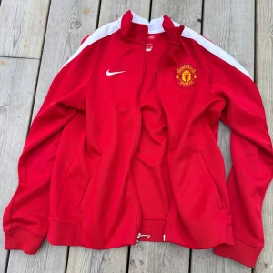 Manchester United röd träningsjacka från Nike - Röd träningsjacka från Nike med Manchester United-logga på bröstet och tryck på ryggen. Jackan har vita ränder längs ärmarna, dragkedja framtill och ståkrage. Perfekt för fotbollsfans som vill visa sitt lagstöd. Satt till acceptabelt skick då små fläckar finns på högern ärm (se kort).
