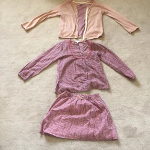 Rosa mönstrad klänning från livly  med matchande kofta - Söt rosa klänning från livly  med småmönstrat tryck och röda detaljer vid halsringningen. Tillhörande ljusrosa kofta med dragkedja. Klänningen har långa ärmar och en stilren look .