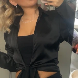 Svart satinskjorta med knyt - Snygg svart skjorta i satin med knyt framtill. Skjortan har lång ärm och en klassisk krage, perfekt för en trendig look. Sitter snyggt och ger en lyxig känsla med sitt blanka material.
