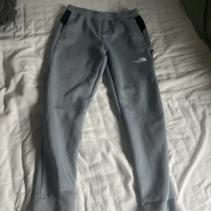 Ljusgrå mjukisbyxor från The North Face - Snygga ljusgrå mjukisbyxor från The North Face med svarta detaljer vid fickorna och logga på låret. Byxorna har elastisk midja och dragkedjefickor på sidorna. Perfekta för chill eller träning.