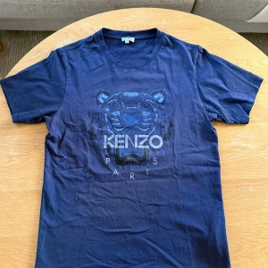 Blå t-shirt från Kenzo med tigertryck - Säljer en blå t-shirt från Kenzo med ikoniskt tigertryck och logga på bröstet. Klassisk passform och rund halsringning. Perfekt för dig som gillar streetwear och vill sticka ut med en snygg detalj från ett känt märke.