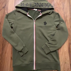 Mörkgrön kofta från Moncler - En grön moncler kofta med text på luvan och fungerande QR kod, dragkedjan lite sliten men syns knappt, inga fläckar och knappt använd, väldigt sällsynt modell från moncler, perfekt passform och håller värmen galant trotts att den är tunn!! Priset går att diskutera!!!