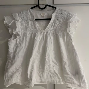 Vit blus med volang från Gina Tricot Young - Säljer en luftig vit topp/blus från Gina Tricot med v-ringning och fina volangdetaljer vid axlarna. Blusen har korta ärmar och dekorativa sömmar upptill. Perfekt topp inför sommarn. Storlek 170. Ny pris 199kr, andvänd fåtal gånger.