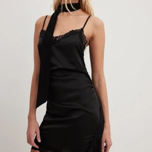 Svart satin klänning med spetsdetaljer - Säljer en svart kort klänning i satin med tunna axelband och vacker spets längs urringning och nederkant. Klänningen har en figurnära passform och en elegant slits framtill. 