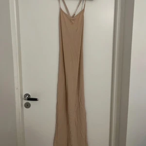 Beige långklänning med smala axelband - Säljer en stilren beige långklänning med smala axelband som korsas i ryggen. Klänningen har en rak passform och är tillverkad i ett mjukt, lätt material som ger en avslappnad look. Perfekt för varma dagar eller sommarens tillställningar.