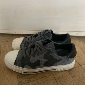 Valentino sneakers med grått camouflage-mönster - Säljer ett par snygga Valentino sneakers med grått camouflage-mönster och vita tåpartier. Skorna har snörning, svart häl och en grov vit sula. Perfekta för dig som vill sticka ut med stilrena detaljer.