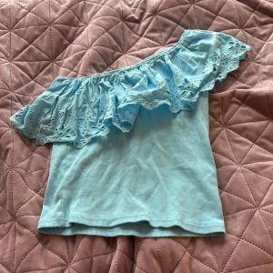 Ljusblå offshoulder topp med volang från Zara - Säljer en ljusblå offshoulder topp från Zara med bred volangkant i broderad spets. Toppen är ribbad och har ett axelband. Perfekt på sommaren när man är tan!🩵