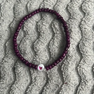 Lila pärlarmband med hjärtdetalj - Sött armband med lila pärlor och en vit pärla med ett rosa hjärta. Perfekt accessoar för att addera lite färg och kärlek till din stil.