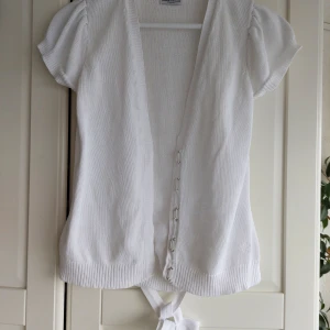 Vit stickad kortärmad kofta från Hunkydory - Säljer en vit stickad kofta från Hunkydory Essentials. Koftan har korta puffärmar, knytband och hyskknäppning framtill. Perfekt för lager-på-lager och enkel att matcha med olika outfits.