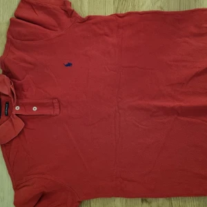 Röd pikétröja från Ralph Lauren - Klassisk röd pikétröja från Ralph Lauren med blå broderad logga på bröstet. Tröjan har korta ärmar, krage och knappar framtill. Perfekt för en stilren och sportig look.
