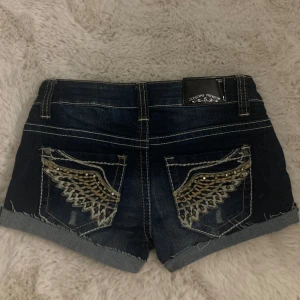 lågmidjade jeansshorts - Snygga mörkblå jeansshorts från ZCOJEANS med uppvikta benslut och dekorativa vingdetaljer i guld och silver på bakfickorna. Klassisk femficksmodell med knapp och dragkedja framtill. Perfekta för sommardagar! Köpt på plick💕