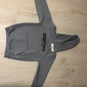 Grå Essentials Fear of God hoodie - Snygg grå hoodie från Essentials Fear of God med svart logga på bröstet och ärmen. Klassisk känguruficka framtill, huva och ribbade muddar. Perfekt för en clean och avslappnad streetstyle-look.