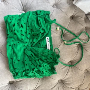 Grön bandeau tubtopp från Zara - Snygg grön bandeau tubtopp från Zara med broderade hål och smockad baksida. Toppen har tunna knytband med pärldetaljer och är perfekt för en färgstark look. Lätt och luftig känsla, passar till sommarens alla outfits.