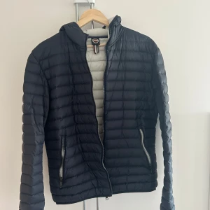 Mörkblå dunjacka från Colmar - Säljer en mörkblå dunjacka från Colmar med horisontella sömmar och huva. Jackan har dragkedja framtill och två sidofickor med dragkedja. Insidan är ljusgrå och jackan har en klassisk, sportig look med Colmar-logga på ärmen.