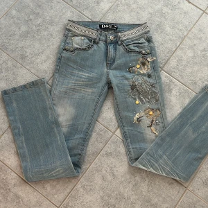Unika Ljusblå lågmidjade jeans - Unika ljusblå jeans från D&N med paljetter och snygga patches på ena benet. Fickor med nitar och bling. Lowwaist med raka/bootcut ben. Perfekta för dig som gillar statement-plagg och detaljer💖Öppen för prisdiskution!