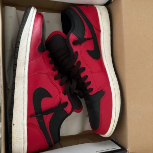 Nike Air Jordan 1 Low SE röd/svart - Säljer ett par Nike Air Jordan 1 Low SE i rött och svart skinn med klassisk svart swoosh och vita sulor. Låga sneakers med svart snörning och ikoniska detaljer. Perfekta för dig som vill sticka ut med en tidlös och sportig look.