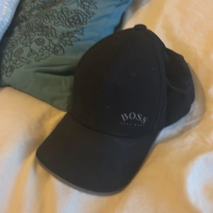 Svart keps från Hugo Boss - Stilren svart keps från Hugo Boss med klassisk böjd skärm och justerbart spänne baktill i metall med Boss-logga. Kepsen har diskret Boss-brodyr framtill och är tillverkad i ett mjukt, bekvämt material som passar perfekt till streetwear eller sportig look. När man har på sig denna då blir man en riktig boss.