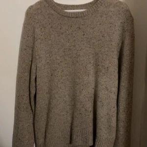 Beige stickad tröja från East West - Beige stickad tröja från East West i storlek S. Tröjan har en klassisk rund halsringning, ribbade muddar och diskreta svarta och bruna prickar i garnet som ger en snygg melerad look. Perfekt för lager-på-lager under kyliga dagar.