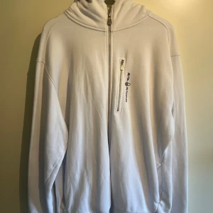 Vit hoodie från Sail Racing - Vit hoodie från Sail Racing i storlek 3XL. Tröjan har huva, half zip-dragkedja och en bröstficka med dragkedja. Broderad logga på bröstet och diskret emblem på axeln. Mjuk bomullskänsla och relaxed fit, perfekt för chill dagar. Köpt för ca 2 år sedan och är i mycket fint skick då endast använd ca 4 gånger p.g.a plagget var för stort för mig 