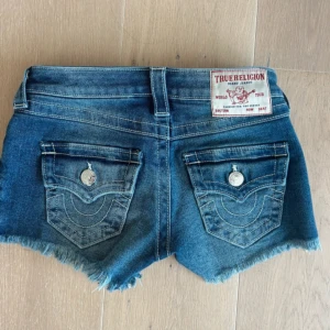 Blå jeansshorts från True Religion - Snygga blå jeansshorts från True Religion med fransig kant och klassiska bakfickor med lock och silverknappar. Shortsen har låg midja och cool slitning framtill. Perfekta för sommaren och ger en avslappnad vibe.