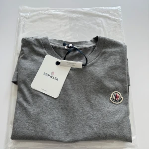 Oanvänd Moncler T-shirt  100 % Bomull.  - Säljer en original Moncler-tshirt från ett exklusivt 3-pack (ord.pris 6 000 kr). Denna är oanvänd och säljs separat – inga kopior. Kvitto saknas, men taggen sitter kvar och tröjan har äkta etikett, materialmärkning och detaljer (se bilder).    Storlek: M Färg: Grå Modell: Klassisk passform