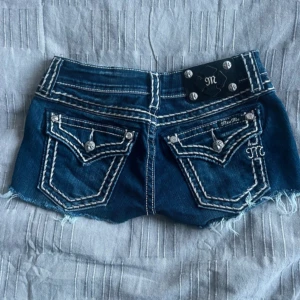 Miss Me jeansshorts med detaljer - Snygga blå jeansshorts från Miss Me med grova kontrastsömmar, slitna kanter och coola bakfickor med dekorativa knappar och broderier. Shortsen har låg midja och klassisk femficksdesign. Perfekta för sommaren när du vill sticka ut lite extra.